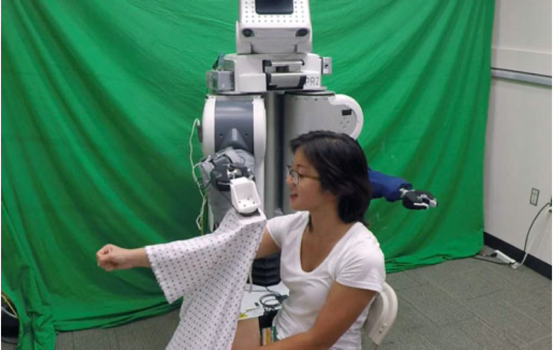 Image to demonstrate robot-assisted dressing. A person sits in front of a robot with their arm outstretched while the robot pulls a cloth gown over the person's arm.